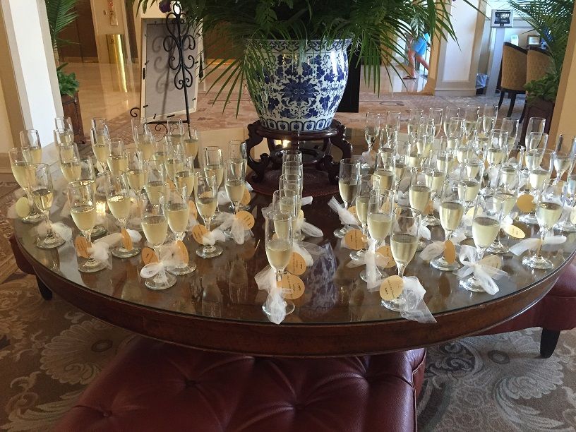 A wooden table with a blue and white vase has a lot of champagne flutes filled halfway with champagne sit around the table. THey have white bows and round tags attached to the stem of the glass.