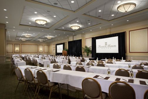 Our Carolina ballrooms has rows of long, rectangular tables et with green and gold chairs face two large projector screens and a stage that has a wooden podium at the front.