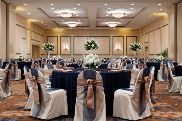 Ballroom of the hotel called the Carolina Ballroom is set with multiple roun tables with blue tablecloths and chairs decorate in blue and mauve ribbons.