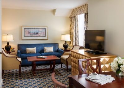 Our Francis Marion Suite features a blue sofa two gold chairs in the living room space. To the right hand side is a small dining table with a few chairs. The space features blue and gold accents throughout.