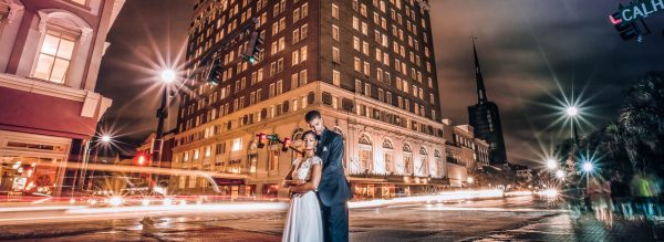 A bride and groom in the center of he photo with the exterior of the hotel behind them. It is night time and the cars headlights stream throughout the streets.
