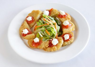 A round white plate with half circle shaped fried green tomatoes with red pepper jelly and a white goat cheese are plated around the edge of the plate. A green salad with shredded carrots and onion sit in a neat pile in the center.