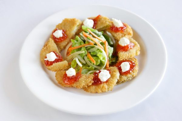 A round white plate with half circle shaped fried green tomatoes with red pepper jelly and a white goat cheese are plated around the edge of the plate. A green salad with shredded carrots and onion sit in a neat pile in the center.