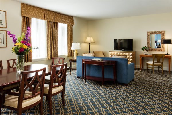 Our Harborview Room Suite is shown with a wooden dining room table with ive chairs, a blue sofa and an orante mirror hangs on teh wall. A desk with one chair is on the right side next to it is a tv. Light shines in through the windows on the left side.