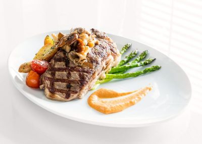 An oval white plate has a large grilled steak on top of roasted potaties and tomatoes. Green asparagus sits under the steak and an orange sauce is neatly smeared in a swoosh to the side.