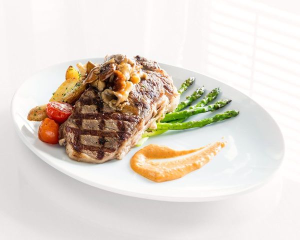 An oval white plate has a large grilled steak on top of roasted potaties and tomatoes. Green asparagus sits under the steak and an orange sauce is neatly smeared in a swoosh to the side.