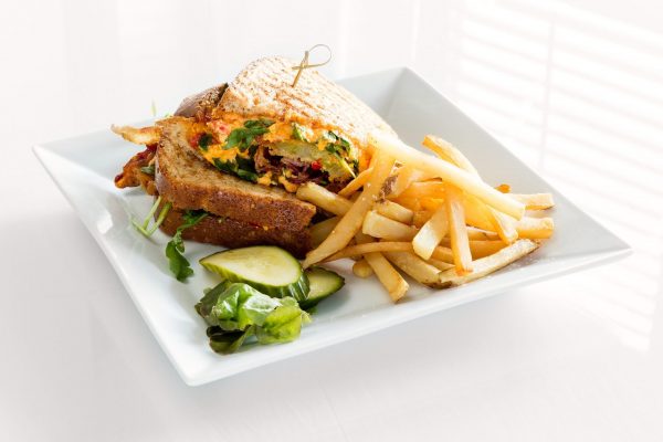A square white plate has a pile of french fires and a triangular cut sandwich with pimento cheese and bacon.