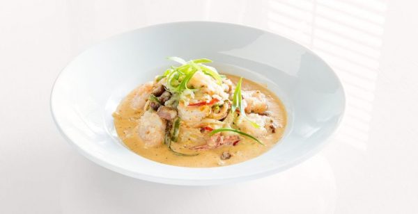 A white bowl is filled with a orange, creamy sauce. Shrimp and red and green peppers with white grits are piled on top of it. Bright green onions sit on the top of the dish.