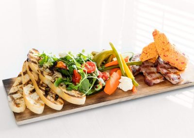 A wooden rectangular board has four slices of grilled bread to the left hand side, a green salad mix with tomatoes and a variety of pickled veggies next to it. Grilled bacon and pimento cheese sit on the end of the board.