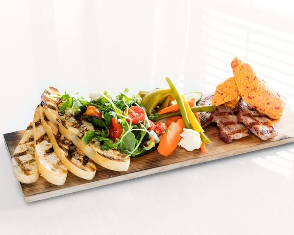 A wooden rectangular board has four slices of grilled bread to the left hand side, a green salad mix with tomatoes and a variety of pickled veggies next to it. Grilled bacon and pimento cheese sit on the end of the board.