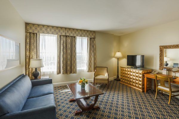 Our downtown view suite shows the living room space that offers a blue sleeper sofa, a wooden coffee table sits in front of it. A desk with a gold chair is to the side with an ornate mirror on the wall.