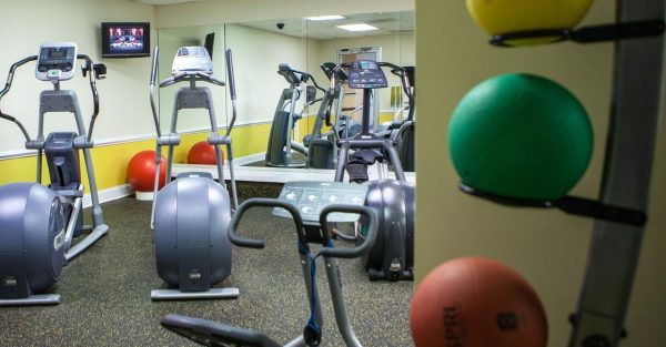 A fitness room with two elliptical machines are shown, a mirror is int he front and small tv sits in the corner. A yellow, red and green weight ball are to the side.