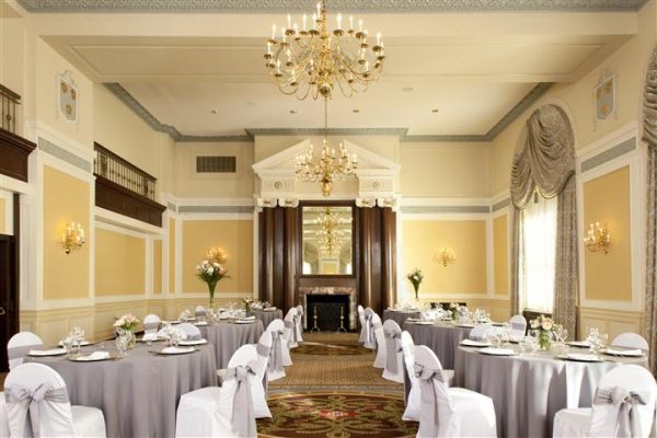 Our colonial ballroom has gorgeous chandeliers that are gold with gold sconces. There are round tables with purple tablecloths and white chairs with purple bows around them.