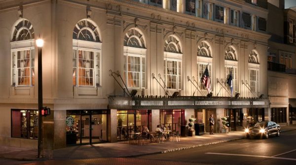 The exterior of the hotel from a corner view. It is evening and a car with its lights on sit in the valet lane to the right side. The building features a few people sitting outside of the coffee shop and the grand windows feature an arch and intricate details in architecture.