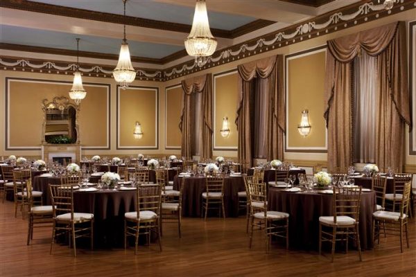 A ballroom in the hotel that is called the Gold Ballroom with wooden floors and gold chairs with antique chandeliers. A grand mirror is against the golden walls.