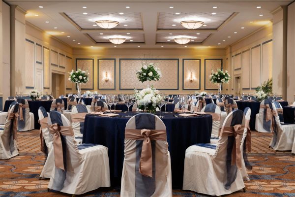 Ballroom of the hotel called the Carolina Ballroom is set with multiple roun tables with blue tablecloths and chairs decorate in blue and mauve ribbons.