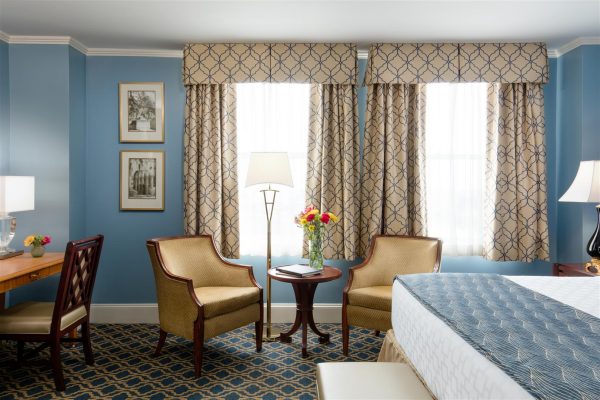 A hotel room with gold and blue accents is features. Two gold chairs sit in front of the windos and a bed is to the right and a small desk is to the left side.
