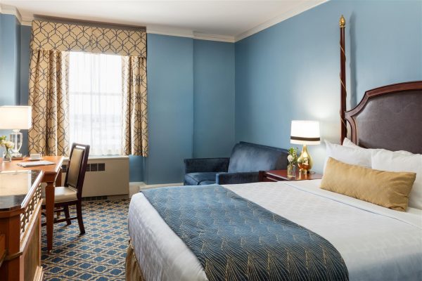 Our traditional room with one queen size bed is featured. Blue and Gold accents are featured throughout. A blie sleeper sofa sits to the right hand side. A small desk is to the left next to a tv console.