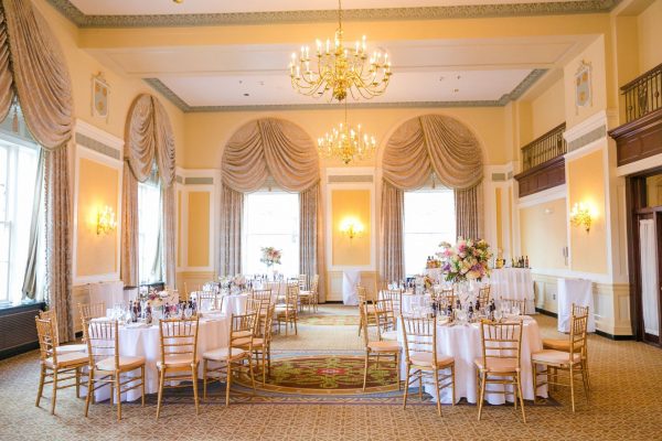 A ballroom space that is brightly lit has six round tables with gold chairs around the, A tall bar is to the back against large windows with elegant curtains. Two antique chandeliers hand from the ceiling.