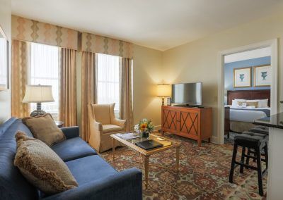 The living room of one of our suites has a blue couch and a yellow chair witha coffee table in the middle. A door leads into the bedroom and shows a neatly made bed.