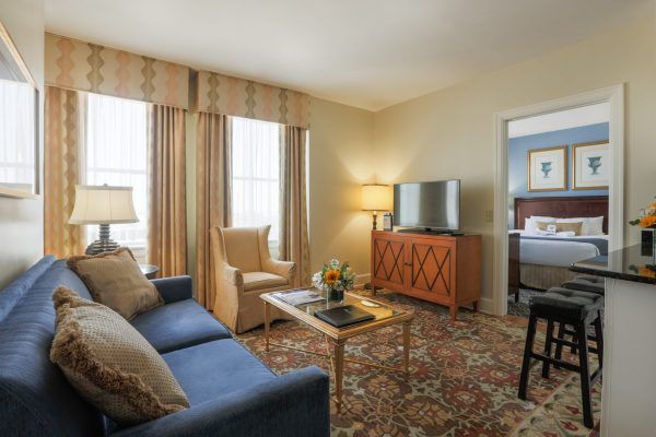 The living room of one of our suites has a blue couch and a yellow chair witha coffee table in the middle. A door leads into the bedroom and shows a neatly made bed.
