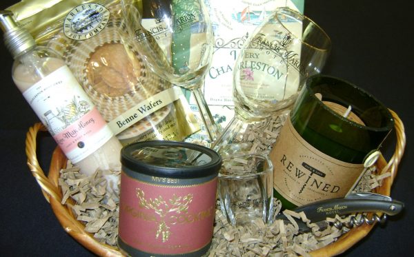 A basket filled with wine glasses, lotion, benne wafers, a book called very charleston, a green candle and a shot glass sit on top of brown paper inside a basket.