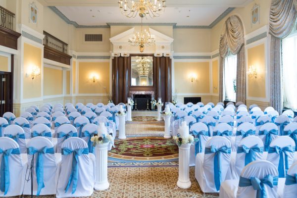 Our Colonial Ballroom is pictured with a wedding ceremony set up. White chairs with light blue bows line featuring the fireplace at hte center of the room. Blue and gold accents are throughout the walls and molding on the ceiling.