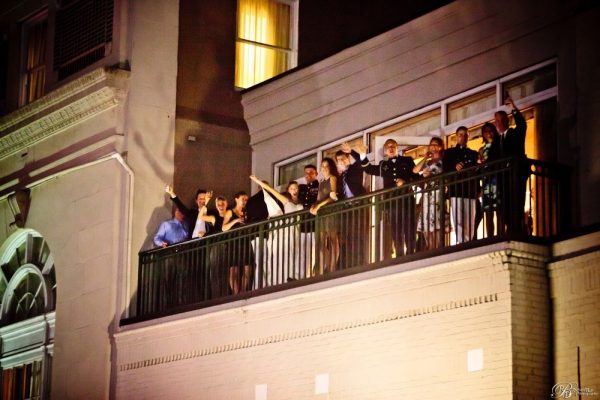 A group of people including a bridge and groom stand out on a small balcony and wave to the camera.