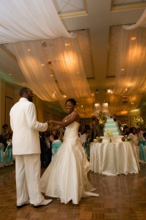 A bride and groom dance together both dressed in all white. Their blue and white wedding cake is placed behind them and their guests