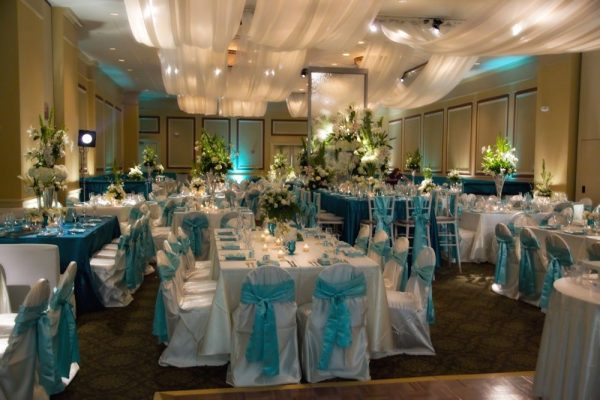 Our Carolina Ballroom during a wedding. Many tables are placed throughout the space with white and blue tableclothes and blue bows around the chairs. White drapery hands from the ceiling and soft lighing is throughout. Large flower centerpieces are spaced throughout each table.