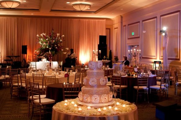 Our Carolina Ballroom during a wedding reception. A cake is in the forefront of the photo with white flower petals around it and candles. In the background are tables with gold chairs. A large flower centeriece with white flowers and candles are throughout. Soft lightiing brightens the darker space. A band sets up in front a curtain in the background.