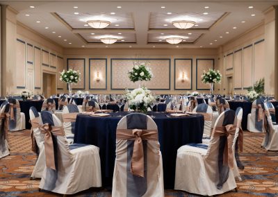 Ballroom of the hotel called the Carolina Ballroom is set with multiple roun tables with blue tablecloths and chairs decorate in blue and mauve ribbons.