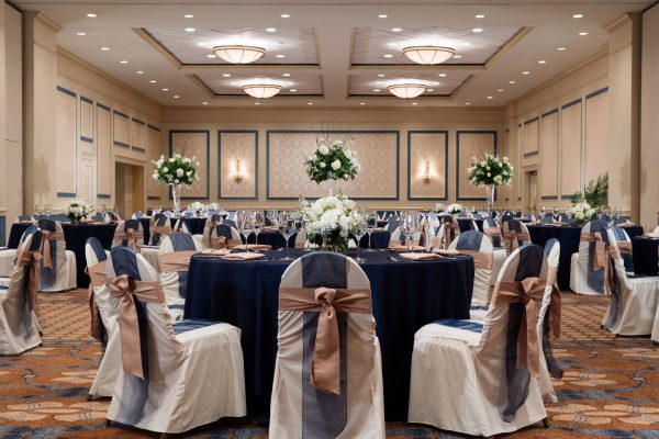 Ballroom of the hotel called the Carolina Ballroom is set with multiple round tables with blue tablecloths and chairs decorate in blue and mauve ribbons.