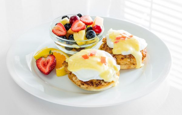 A round plate has two poached eggs with a yellow hollandaise sauce over top of them. A small clear bowl has a fruit salad next to the eggs.