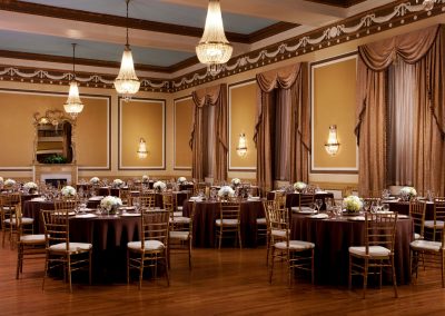 A ballroom in the hotel that is called the Gold Ballroom with wooden floors and gold chairs with antique chandeliers. A grand mirror is against the golden walls.