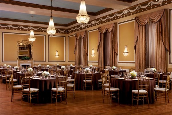 A ballroom in the hotel that is called the Gold Ballroom with wooden floors and gold chairs with antique chandeliers. A grand mirror is against the golden walls.