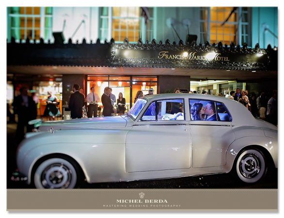 A bride and groom kiss in the backseat of an antique white car outside the hotel at nighttime.