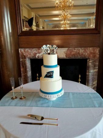 A round table with a light blue runner has a three tiered white and blue wedding cake. Two empty champagne flutes sit to the side.