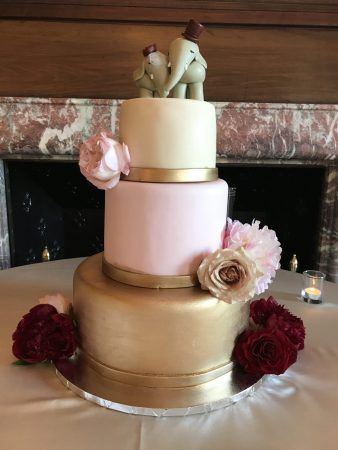 A three tiered wedding cake that is gold, pink and cream color has two elepahnts on the top of the cake. Pink and red roses sit around the edge of the tiers.