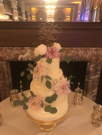A white four tiered wedding cake has light pueple flowers around it and a cake topper reads love you more sits on the very top.