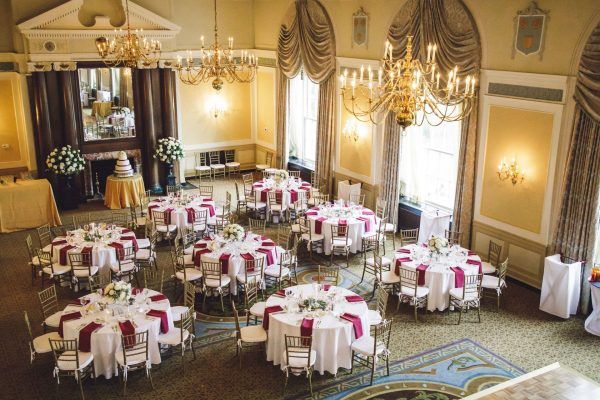 An aerial photo of the colonial ballroom shows multiple round tables with red naplins and gold chairs. A three tiered wedding cakes sits on a small round table in front of an ornate fireplace. Antique chandeliers hand from the ceiling.