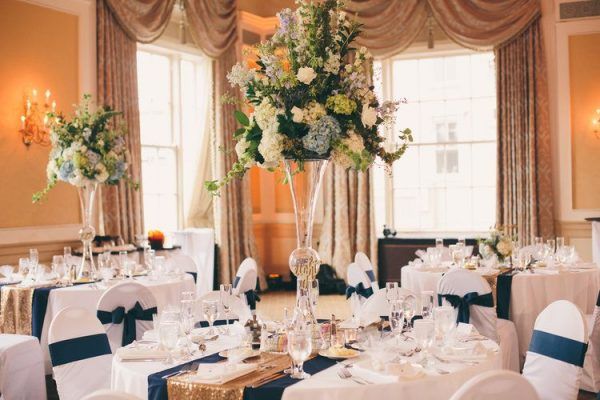 A large floral centerpiece sits in the center of a round table with a white tablecloth and blue and gold table runnder.