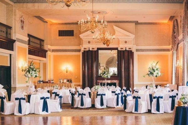 Our colonial ballroom has a dance floor in the center and multiple round tables with blue and white accents. Large floral centerpieces sit in the middle of three tables and a long table set up to be a bar sits in the back left hand side of the room.