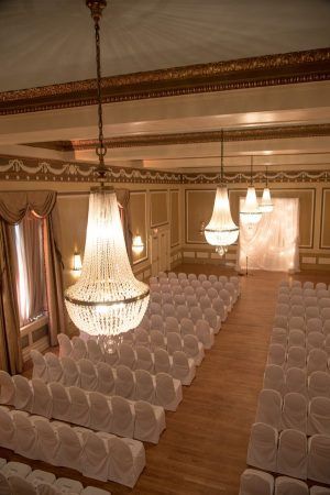 A view of our gold ballroom from above shows the antique chandeliers close up and below are many rows fo chairs covered in white chair covers lined up in rows for a wedding ceremony.