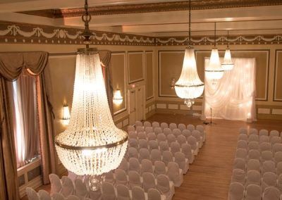 A view of our gold ballroom from above shows the antique chandeliers close up and below are many rows fo chairs covered in white chair covers lined up in rows for a wedding ceremony.