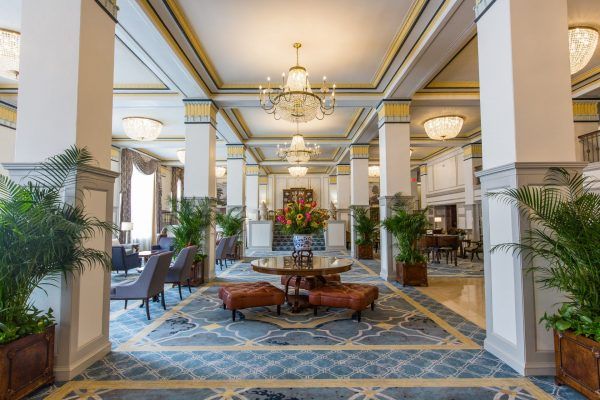 The lobby of the hotel showcases plenty of natural light with blue and gold carpet. A beautiful flower display sits in the center of the room on a round table.