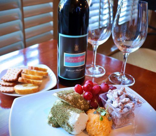 A plate with cheeses and grapes. A bottle of wine and two wine glasses site behind the plate.