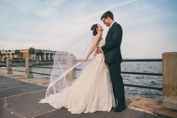 A bride and groom stand facing each other as the bride's veil flows in the wind. They are standing next to the harbor.
