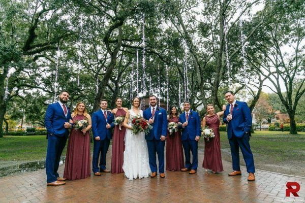 A bridal party stands together with the bride and groom in the center. Trees are behind them with hanging string lights are above the,.