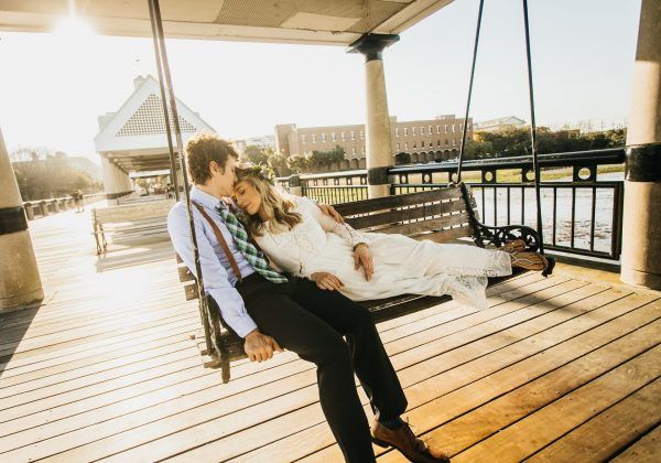 A bride and groom sit with each other on a park swing bench. The bride lays against the groom's chest.